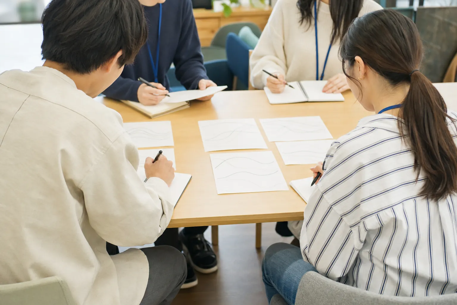 月例研修の様子。資料を囲んでメモを取る学生講師たちの手元と後ろ姿（顔が写らない構図）。テーブルに研修資料が並ぶ
